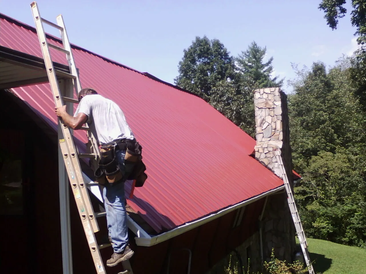 Roofer on a ladder beside a red metal roof and stone chimney — Soffit & Fascia Repair by Roofing Brothers in Taftville